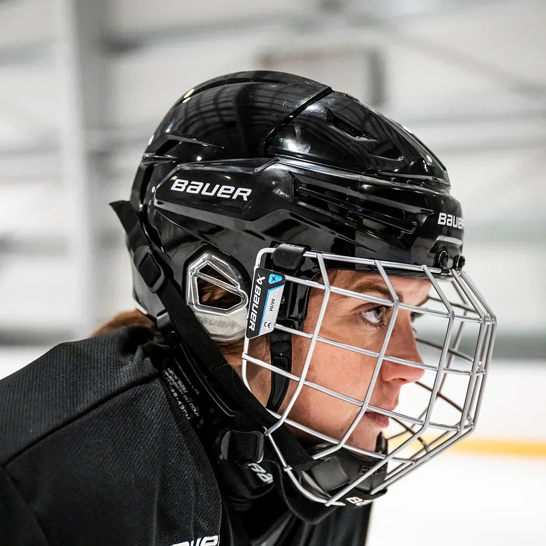 A photo of the The Bauer RE-AKT 70 hockey helmet being worn by a woman athlete wearing a black jersey, in a hockey arena.