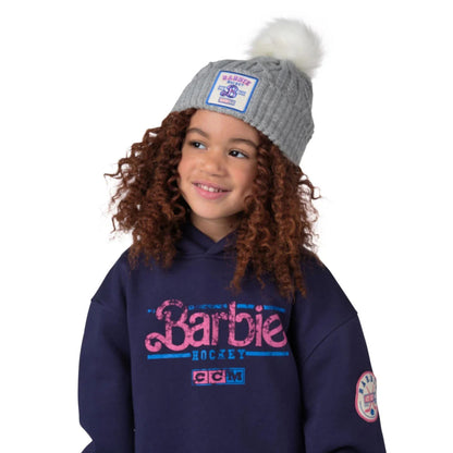 A photo of a child wearing a navy Barbie hoodie sweater and a grey Barbie™ x CCM Grey Pom Knit Youth Toque.